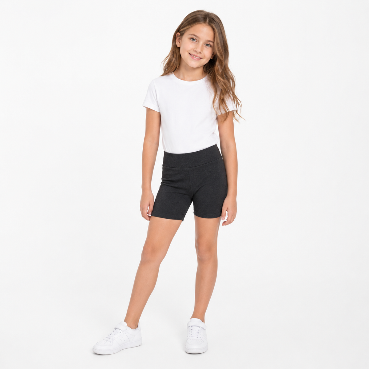 Niña con polera blanca, calza corta escolar negra y zapatillas blancas en vista frontal